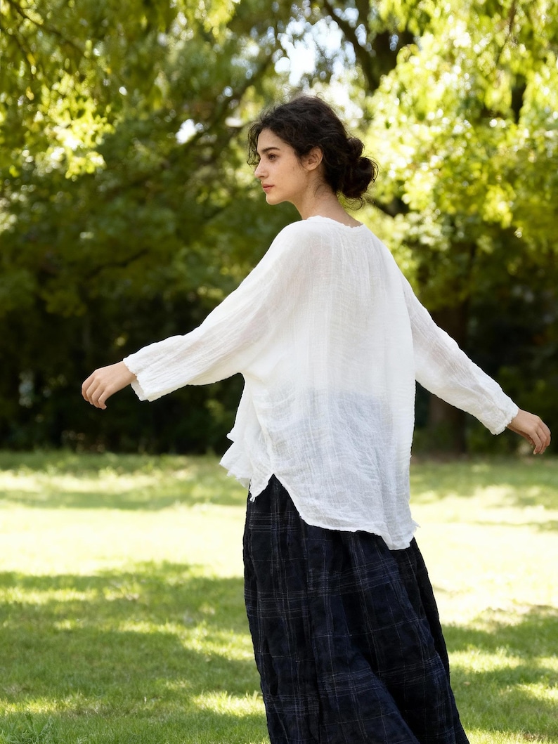 May include: A white, long-sleeved blouse with a loose fit and a scoop neckline. The blouse is paired with a dark blue plaid skirt. The image is taken outdoors with a green background.