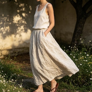 May include: A two-piece linen outfit in a neutral color. The top is a sleeveless tank, and the skirt is a flowing midi-length with pockets. The outfit is worn with brown sandals. The background includes a tree and flowers.