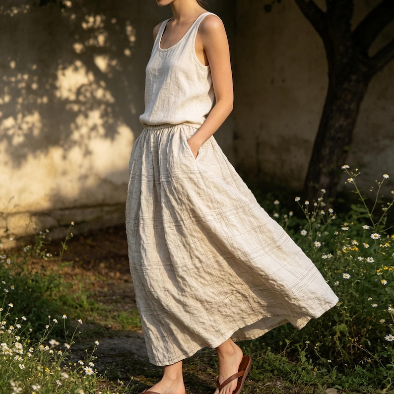 May include: A two-piece linen outfit in a neutral color. The top is a sleeveless tank, and the skirt is a flowing midi-length with pockets. The outfit is worn with brown sandals. The background includes a tree and flowers.