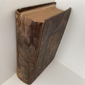 Puede incluir: Un libro antiguo con una cubierta de cuero marrón desgastada. Las páginas están envejecidas, con un color marrón claro. La cubierta muestra signos de desgaste, lo que sugiere que es una antigüedad o un artículo muy usado. El libro está inclinado.