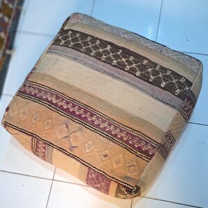 Peut inclure: Un coussin de sol carré doté d'une housse en textile tissé avec des rayures horizontales dans des tons beige, brun clair, marron et violet sourd. Le tissu présente un motif texturé avec des détails géométriques et des bordures.