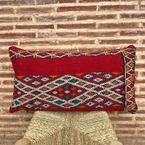 May include: Rectangular red pillow with a geometric pattern in white, orange, green, and black. The pillow has a solid red top and a patterned bottom. The pillow is resting on a woven surface.