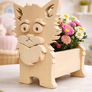 Può includere: Una fioriera in legno a forma di gatto con finitura in legno chiaro. Il gatto ha grandi occhi, una barba e una scatola rettangolare per i fiori. La fioriera è piena di fiori rosa, bianchi e gialli.