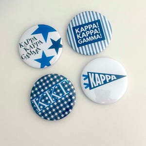 May include: Four round buttons with blue and white designs. One button has blue stars and the text "KAPPA KAPPA GAMMA". Another has blue stripes and the text "KAPPA! KAPPA! GAMMA!". A third button has a blue gingham pattern with the letters "KKΓ". The last button has a blue pennant with the word "KAPPA".