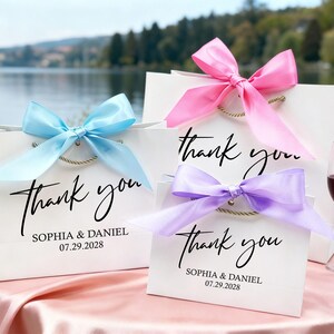 May include: White gift bags with "Thank you" in elegant script, tied with light blue, pink, and purple satin bows. Each bag includes the names "Sophia & Daniel" and the date "07.29.2028".