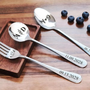 May include: A set of stainless steel cutlery, including a fork and two spoons, with engraved initials and dates. The spoon and fork are on a wooden tray. The spoon has the initials "A & D" and the dates "06.23.2028", "11.09.2028", and "09.13.2028" are engraved on the handles.