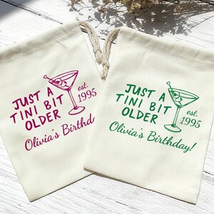 May include: Two off-white drawstring bags with text and a martini glass illustration. One bag has purple text that reads "JUST A TINI BIT OLDER Olivia's Birthd est. 1995". The other bag has green text that reads "JUST A TINI BIT OLDER Olivia's Birthday! est. 1995".
