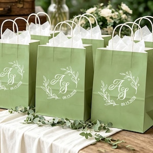 Può includere: Una collezione di sacchetti regalo in carta verde salvia con manici in carta intrecciata bianca e inserti in carta velina bianca. Ogni sacchetto presenta un monogramma bianco stampato con le lettere AS circondate da una corona botanica e la data 09.15.2028.