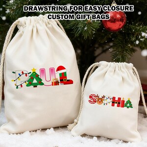 May include: Two cream-coloured drawstring gift bags with festive embroidery. One bag says "PAUL" with a Christmas tree, lights, and Santa boot. The other says "SOPHIA" with a reindeer, lights, and tree. Text at the top reads "DRAWSTRING FOR EASY CLOSURE CUSTOM GIFT BAGS."