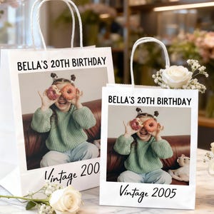 Pu&ograve; includere: Due sacchetti regalo di carta bianca con manici bianchi. Ogni sacchetto presenta una foto di una giovane persona che tiene delle ciambelle sugli occhi. Il testo sui sacchetti recita "BELLA'S 20TH BIRTHDAY" e "Vintage 2005" e "Vintage 200".