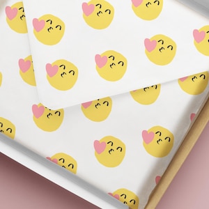 May include: White tissue paper with a repeating pattern of yellow emoji faces with pink hearts. The tissue paper is inside a cardboard box with a white interior and a light brown exterior. The background is a solid pink color.