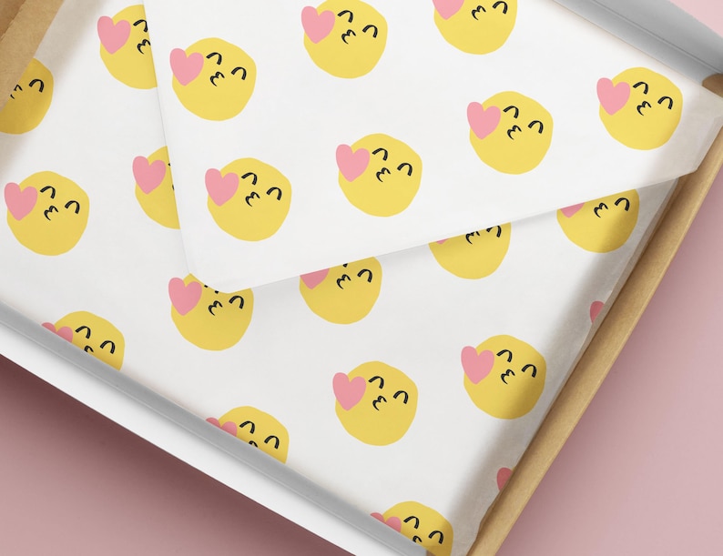 May include: White tissue paper with a repeating pattern of yellow emoji faces with pink hearts. The tissue paper is inside a cardboard box with a white interior and a light brown exterior. The background is a solid pink color.