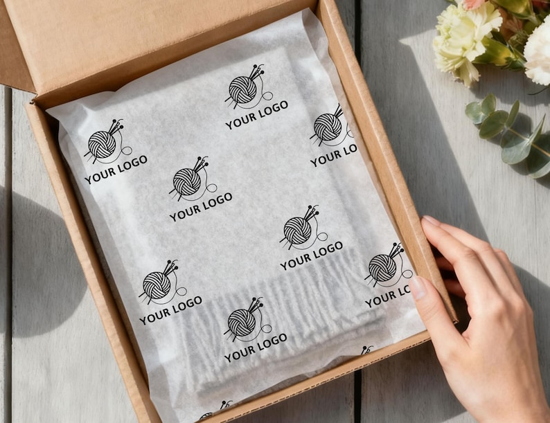 May include: A cardboard box with tissue paper featuring a repeating pattern of a yarn ball and knitting needles with the text "YOUR LOGO". The box is open, revealing the contents, and a hand is visible.
