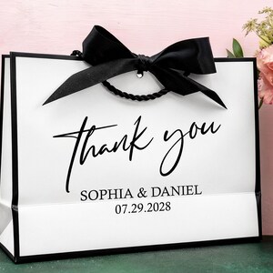 May include: A white gift bag with black edging and a black bow. The words "thank you" are written in elegant script, with the names "SOPHIA & DANIEL" and the date "07.29.2028" below. The bag has a twisted black rope handle.