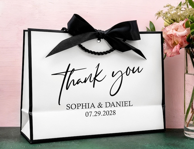 May include: A white gift bag with black edging and a black bow. The words "thank you" are written in elegant script, with the names "SOPHIA & DANIEL" and the date "07.29.2028" below. The bag has a twisted black rope handle.