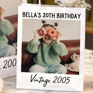 Pu&ograve; includere: Un sacchetto regalo bianco con una foto di una giovane persona che tiene delle ciambelle sugli occhi. Il sacchetto presenta la scritta "BELLA'S 20TH BIRTHDAY" e "Vintage 2005" in corsivo nero.