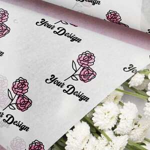 May include: White tissue paper with a repeating pattern of pink roses and the text "Your Design" in black script. A bundle of white flowers is in the lower right corner. The paper is folded over a light purple surface.
