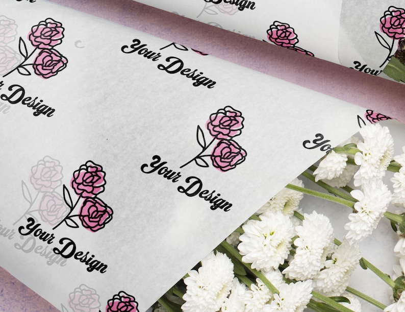 May include: White tissue paper with a repeating pattern of pink roses and the text "Your Design" in black script. A bundle of white flowers is in the lower right corner. The paper is folded over a light purple surface.