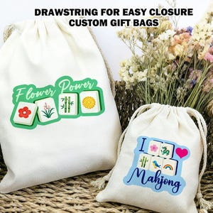 May include: Two white drawstring gift bags with Mahjong tile designs. One bag features "Flower Power" with tile illustrations. The other bag says "I Love Mahjong" with tile illustrations, a heart, and a rainbow. The bags are in a woven basket.