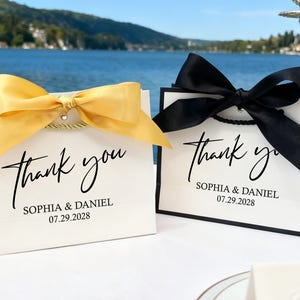 May include: Two white gift bags with black trim, each with a large satin bow. One bow is gold, the other black. The bags read "thank you" with a name and date, ideal for wedding favours or special events.
