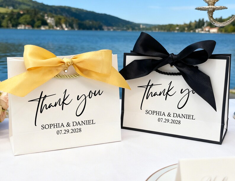 May include: Two white gift bags with black trim, each with a large satin bow. One bow is gold, the other black. The bags read "thank you" with a name and date, ideal for wedding favours or special events.