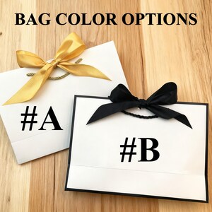 May include: Two white gift bags with black text and bows. One bag has a gold bow and the text "#A". The other bag has a black bow and the text "#B". The words "BAG COLOR OPTIONS" are at the top.