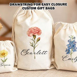 以下が含まれることがあります： クリーム色の巾着ギフトバッグ3つ。デイジー、ピンクのカーネーション、青いデルフィニウムの花柄。各バッグには花の下に名前が印刷されています：Aurora、Scarlett、Emily。「DRAWSTRING FOR EASY CLOSURE CUSTOM GIFT BAGS」の文字が上部にあります。