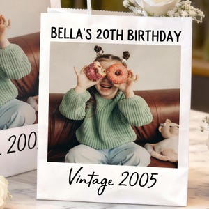 Pu&ograve; includere: Un sacchetto regalo bianco con una foto di una giovane persona che tiene delle ciambelle sugli occhi. Il sacchetto presenta la scritta "BELLA'S 20TH BIRTHDAY" e "Vintage 2005" in nero.