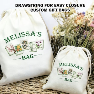 May include: Two off-white drawstring gift bags with the text "MELISSA'S BAG" in green, along with floral and dragon tile illustrations. The bags are in a woven basket with dried flowers. The text "DRAWSTRING FOR EASY CLOSURE CUSTOM GIFT BAGS" is at the top.