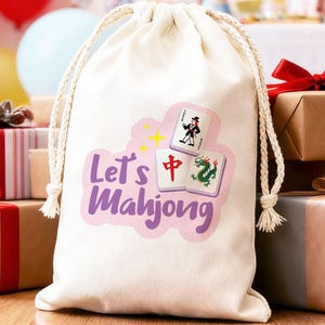May include: A cream-coloured drawstring bag featuring the text "Let's Mahjong" in purple, and Mahjong tile illustrations. The bag is set amongst wrapped gifts and balloons, implying a party or celebration.
