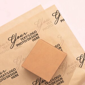 May include: A square, light brown box rests on top of two sheets of light brown paper. The paper has a repeating pattern of the word "Your" in a script font, along with the text "TEXT/LOGO PHOTO/DESIGN HERE".