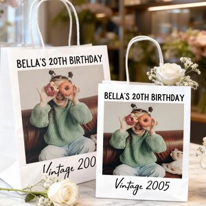 Pu&ograve; includere: Due sacchetti regalo di carta bianca con manici bianchi. Ogni sacchetto presenta una foto di una giovane persona che tiene delle ciambelle sugli occhi. Il testo sui sacchetti recita "BELLA'S 20TH BIRTHDAY" e "Vintage 2005" e "Vintage 200".