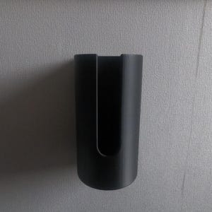 Può includere: Un supporto nero a forma di U, forse per un accessorio per aspirapolvere, è montato su una parete grigio chiaro strutturata. Il supporto ha una base arrotondata e una fessura verticale al centro.