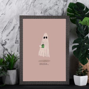 May include: A framed art print featuring a whimsical ghost illustration. The ghost, with black eyes, holds a green mug and is draped in a white sheet. The print has a pink background with the word "Shhhh." in black.