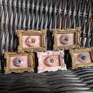 May include: Five small rectangular frames containing sculpted human-like eyes with realistic skin texture. Four frames are ornate gold, while one central frame is white with red speckles. Each eye features a distinct iris colour: brown, green, blue, and yellow.