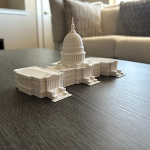 May include: A white miniature replica of the United States Capitol Building. The model features detailed architectural elements, including a domed roof and columns. The building is placed on a dark gray surface.