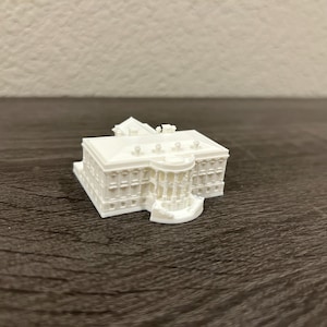 May include: A small, white miniature of the White House. The model features detailed architectural elements, including columns, windows, and the building's distinctive facade. The miniature is set on a dark wooden surface.