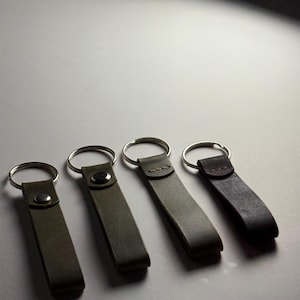 May include: Four leather keychains in varying shades of green and brown. Each keychain features a rectangular leather strap attached to a silver metal ring. The keychains are arranged in a row, showcasing their different colors and textures.