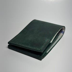 May include: A dark green leather wallet with red stitching. The wallet is closed, with a slight view of the interior. The leather has a textured appearance. The wallet is likely designed to hold cards and cash.