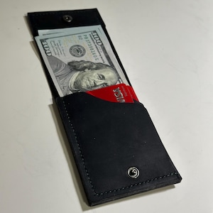 Può includere: Portafoglio in pelle nera con chiusura a scatto, contenente una pila di banconote da cento dollari e una carta di credito rossa. Il portafoglio è rettangolare e ha un design minimalista, con cuciture visibili.