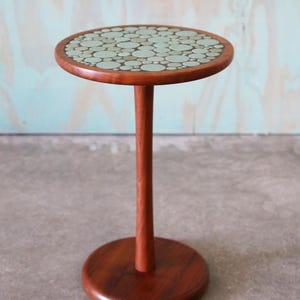 May include: A small, round side table with a dark wood finish. The tabletop features a mosaic design of light green and off-white circles. The table stands 28 inches tall and is supported by a single, slender leg and a circular base.