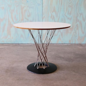 May include: A modern side table with a white circular top and a unique wire base. The base is made of thin, intertwined metal rods that converge at a black circular base. The table's design is minimalist and stylish.