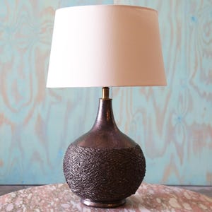 May include: A table lamp featuring a dark brown, textured, spherical base with a cross-hatched pattern and a smooth, tapered neck. The lamp is topped with a simple, off-white, cylindrical fabric lampshade and includes a brass-colored metal fitting.