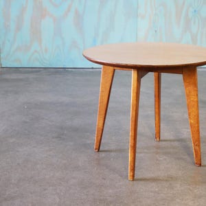 May include: A small, round wooden table with four angled legs. The tabletop is light brown, and the legs are a lighter shade of brown. The table is approximately 24 inches in diameter and 20 inches tall.