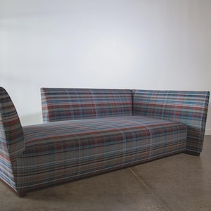 May include: A mid-century modern sofa with a plaid pattern in shades of blue, red, and gray. The sofa has a low profile with a curved arm on one side and a straight back. The upholstery is a woven fabric.