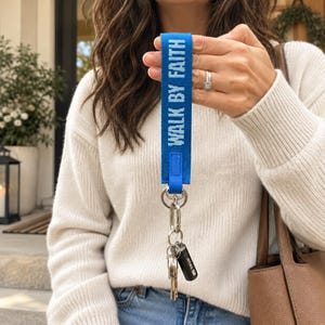 May include: A bright blue fabric wristlet keychain featuring the white embroidered text WALK BY FAITH. The strap is attached to a silver-toned metal split ring and a lobster clasp, which holds a collection of silver keys and a small black cylinder.