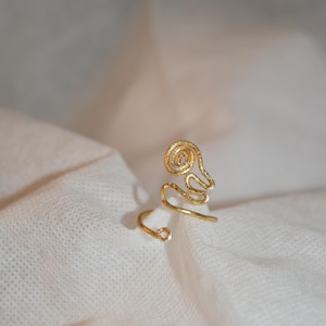 May include: A gold-colored ring with a unique, abstract design. The ring features a swirling, spiral-shaped top and a delicate band that wraps around the finger. The ring is made of a textured metal and sits on a white fabric.