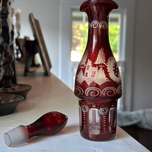May include: A ruby red glass decanter with a matching stopper. The decanter features white etched designs of a village scene and decorative swirls. The stopper has a clear glass tip. The decanter is displayed on a white surface.