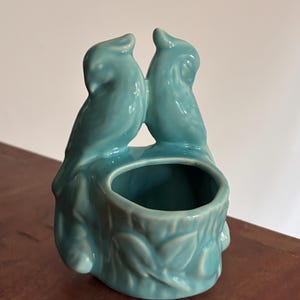 May include: A glossy turquoise ceramic planter featuring two birds perched atop a textured tree stump base. The birds are positioned facing each other, and the cylindrical opening is integrated into the center of the stump design.