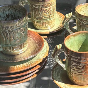 May include: A set of ceramic mugs and saucers featuring a reactive glaze in shades of sage green and earthy brown. Each piece displays a raised, textured relief pattern with geometric and organic motifs. The mugs have sturdy, sculpted handles.